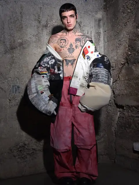 Shirtless tattooed model in a patchwork newspaper-print jacket and pink trousers against a concrete wall