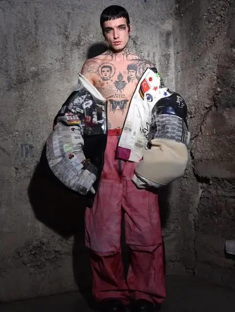 Shirtless tattooed model in a patchwork newspaper-print jacket and pink trousers against a concrete wall