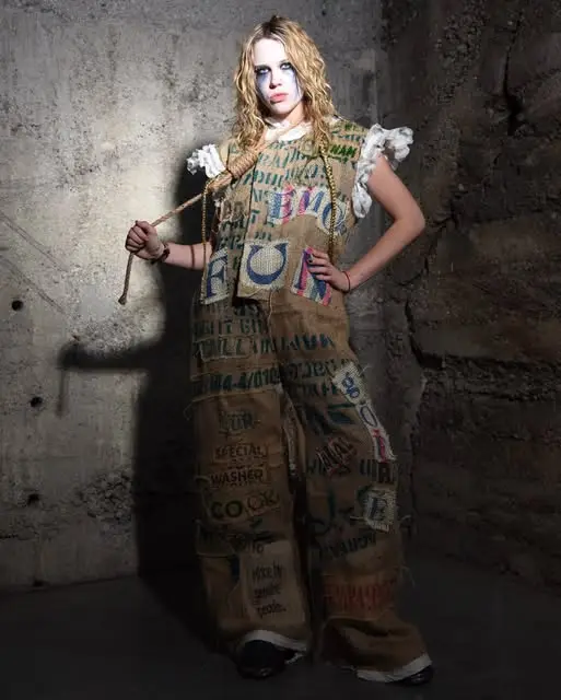 Model with white face paint in a hand-printed sackcloth suit with a stick, concrete wall