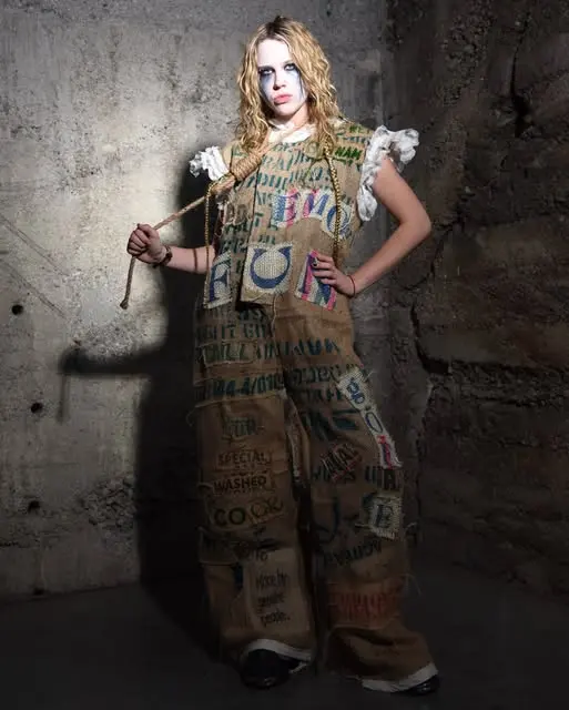 Model with white face paint in a hand-printed sackcloth suit with a stick, concrete wall