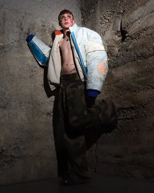 Model in a light patchwork jacket and camouflage trousers leaning against a concrete wall