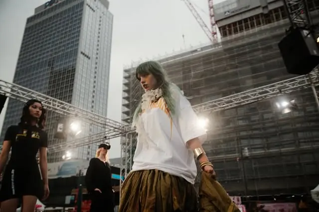 Green-haired model walking an outdoor runway in a white tee and olive parachute skirt