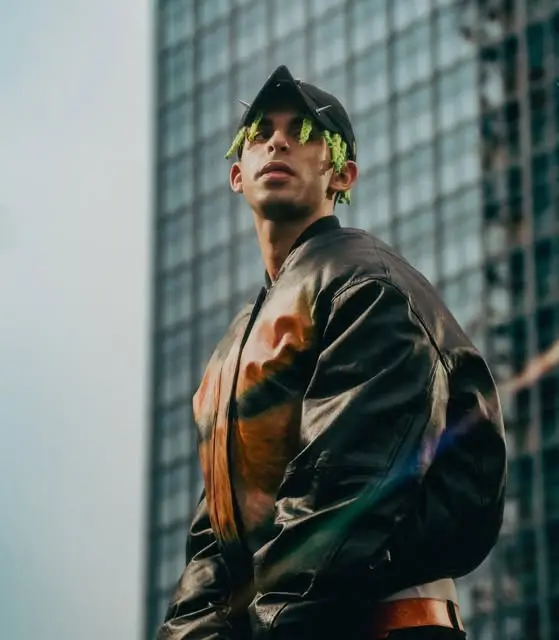 Model in a dark leather-look bomber jacket and cap in front of a glass high-rise