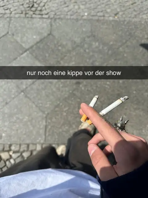 Snapshot of a hand with a cigarette on the pavement, captioned “nur noch eine kippe vor der show”